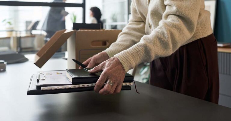 Senior Caucasian woman packing personal belongings and office supplies into cardboard box on desk