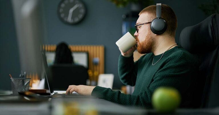Man wearing headphones while working on laptop, holding a coffee cup. Office setting with blurred background including clock and framed picture