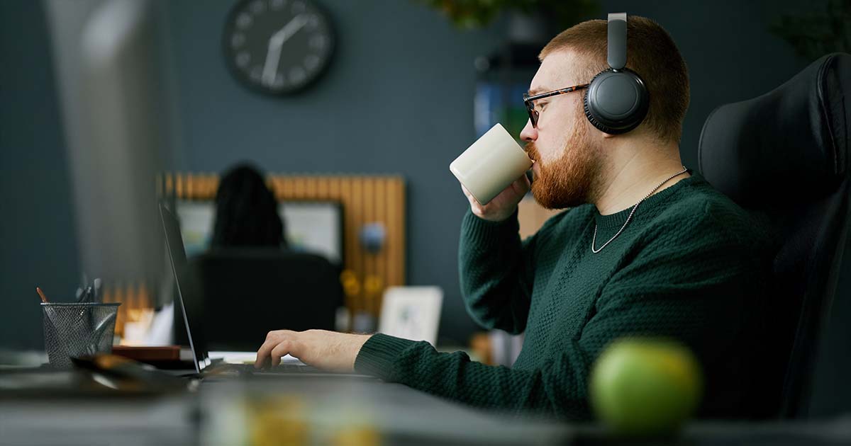 Man wearing headphones while working on laptop, holding a coffee cup. Office setting with blurred background including clock and framed picture