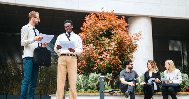 Business people gather in an outdoor urban setting to hold a discussion and review documents, fostering collaboration and communication, surrounded by greenery and a modern architectural backdrop.