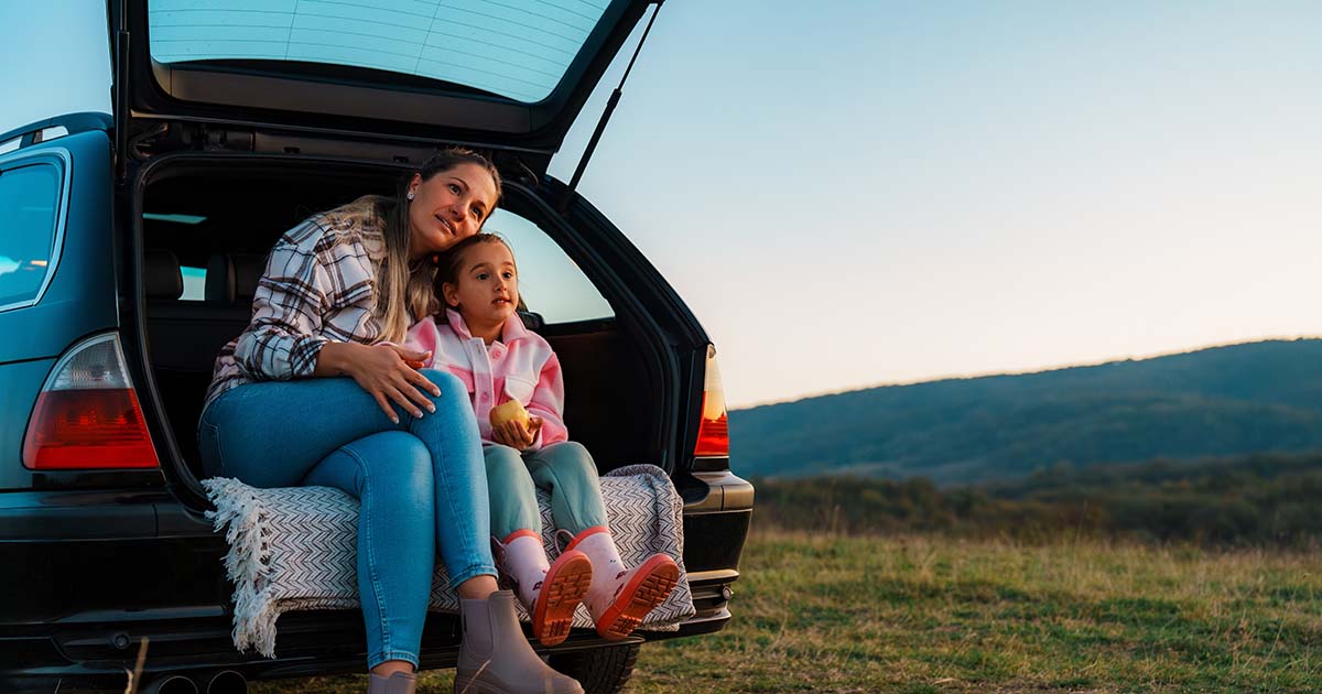 Mother and daughter are sitting in the open trunk of their car, embracing each other and enjoying a peaceful moment while admiring a beautiful mountain landscape during a road trip