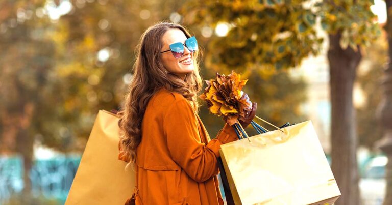 happy stylish woman in orange trench coat with shopping bags and autumn yellow leaves in the city.