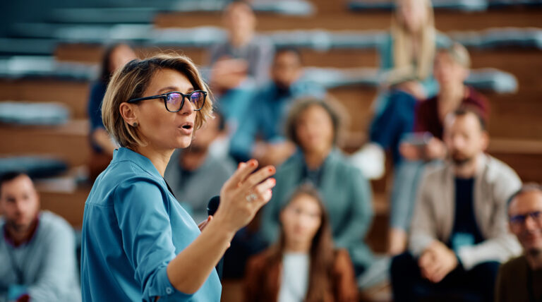 woman with glasses speaking to group of professionals in an auditorium