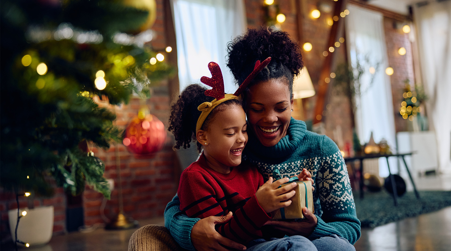 festive mom holding daughter wearing antler headband opening gifts